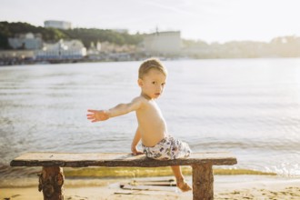Boy child sitting on a wooden bench with his back on the beach near the water and showing his hand