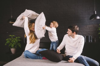 Parents with smiling child playing pillow fight on bed, happy family having fun in bedroom,