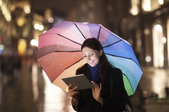 Happy young girl with colorful umbrella using tablet computer outdoor. She standing in the city