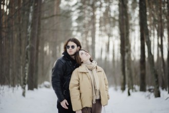 Couple in a snowy forest with the woman leaning on the man, both in winter attire among towering