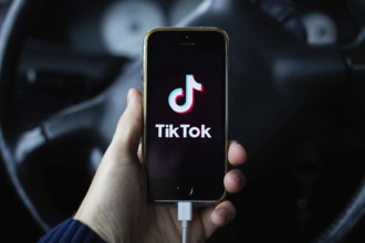 A hand holding a smartphone with TikTok app in front of a steering wheel inside a car
