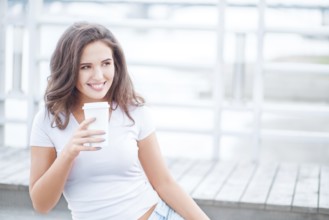Beautiful young woman holding takeaway cup of coffee and sitting on bridge against water background
