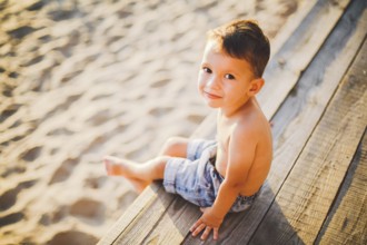 Little Caucasian boy child sitting on wooden pier sandy beach, summer time, sea holiday near water.