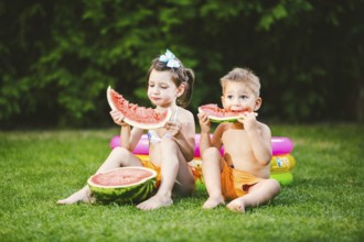Funny little kids brother and sister eating watermelon on green graß near inflatable pool in yard