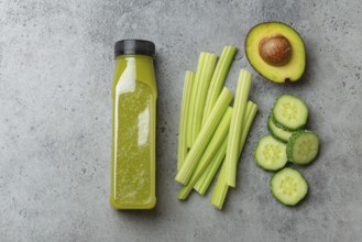 Bottle of green vegetable smoothie with fresh celery sticks, cut avocado, cucumber on gray concrete