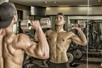 Handsome shirtless muscular young man exercising shoulders in gym with dumbbells