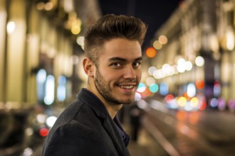 Attractive young man portrait at night with city lights behind him, smiling and looking at camera