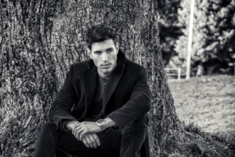 Handsome young man leaning against tree, looking to a side, in a sunny day wearing a black coat