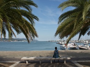 Woman sitting on a bench under palm trees while looking at the sea and the harbour with boats, La