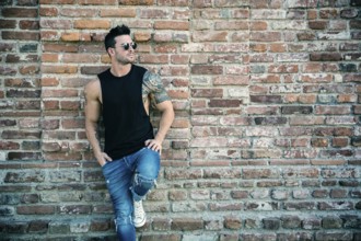 Attractive muscular man standing outdoor leaning against old brick wall, looking to a side