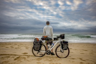 A cycle packer long distance cyclist stand in front of his bicycle on the beach while looking at