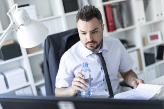 A man is sitting at a table in the office, working with documents and holding a bottle of water in