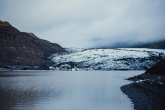 Expansive glacial view with mountains towering over a vast cold lake under a cloudy sky, iceberg,