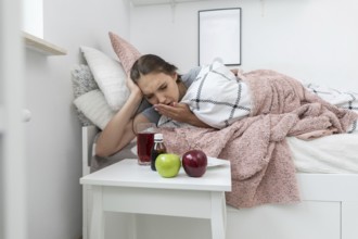 On the bed lies a teenager and in front of her stands a table with apples and medicines that must