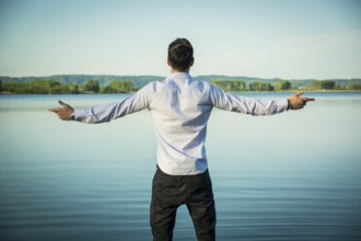 Young man in shirt, outdoor with arms spread open enjoying freedom in front of lake, seen from the