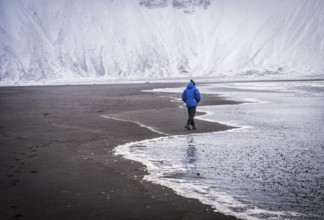 Full body back view of unrecognizable male explorer in blue warm jacket walking along waving sea