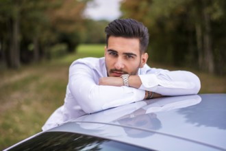 Portrait of young attractive man in business suit leaning on his new stylish car outdoor in
