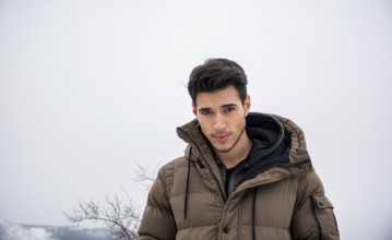Handsome man in outerwear sitting while looking at camera. Snowy landscape on background