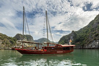 Traditional wooden boat sails on turquoise water between green hills under a cloudy sky, sailing