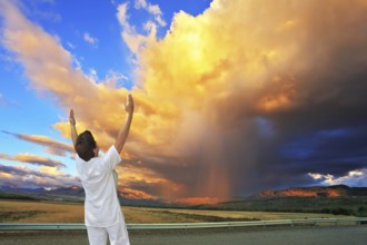 Giant cloud illuminated by the sunset. Yoga on the road. Elderly woman in white performs asana Sun