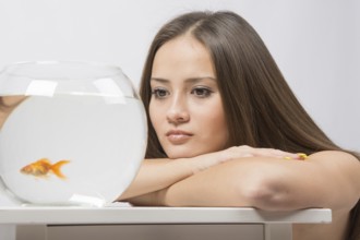 A young girl sits next to a round aquarium in which swimming goldfish
