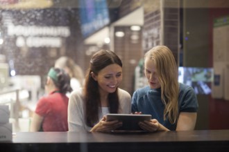 Smiling blonde and brunette women have fun in the restaurant while watching something on the tablet