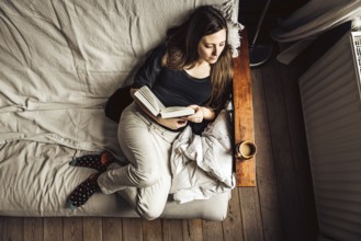 Hig angle view of a 30 yo woman in her sofa, reading a book with a coffee, Brussels