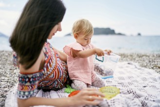 Mom lies on a blanket on a pebble beach next to a sitting little girl.