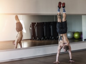 Back view of tattooed shirtless man doing handstand in sportive gym against mirror