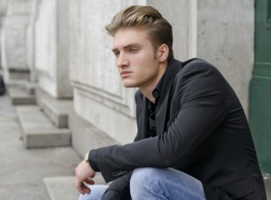 Attractive blond young man in jeans and jacket, sitting outdoors looking off camera