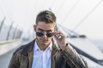 Handsome trendy young man standing on a sidewalk, wearing sunglasses and a fashionable leather