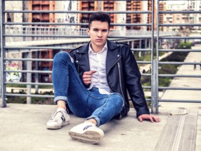 One handsome young man in urban setting in European city, sitting, wearing black leather jacket and