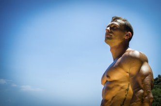 Handsome young muscle man at the seaside, outdoors, showing muscular pecs and torso in front of the