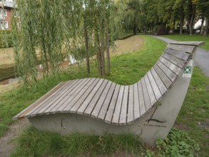 A curved wooden bench stands under a willow tree along a park path, suitable for relaxation,