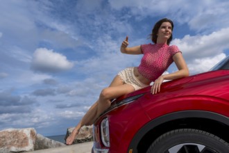 A gorgeous redhead model sits confidently on the hood of her car, soaking in the summer breeze as