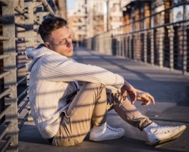 Handsome trendy young man, sitting in city setting at sunset wearing a fashionable sweater or