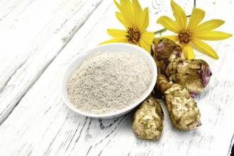 Flour of Jerusalem artichoke in a white bowl with yellow flowers and vegetables on the background