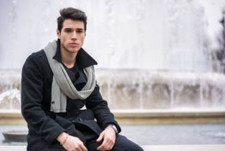Stylish trendy young man sitting outdoor in front of big fountain looking at camera