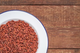 Dried goji berries on white metal enamel plate against rustic wood, top view with a copy space