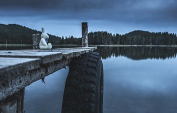 Fluffy toy bunny sitting on a pier. Shore of a mountain lake