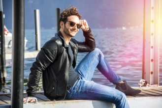 Handsome man on a lake in a sunny, peaceful day, sitting on a wood pier, thinking or meditating