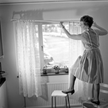 1960s home decorating. A woman at home putting up the curtains in the room window. All by herself