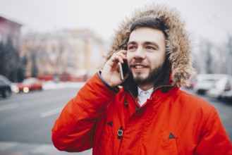 Young guy with beard and red jacket in hood a student uses mobile phone, holds in his hand near the