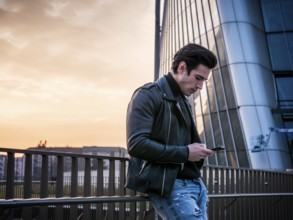 One handsome young man in urban setting in modern city, standing, wearing black leather jacket and