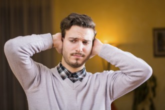 Handsome young man covering his ears, stressed or unhappy because of too much noise. Indoors shot