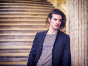 A young, long-haired attractive man standing next to a marble column, leaning onto it, wearing