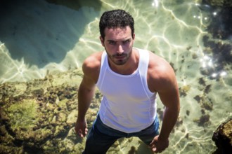Attractive young athletic man in the sea or ocean by the rocky shore, wearing wet t-shirt, serious