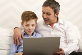 Father and son sitting on the sofa at home. Happy dad embracing boy and watching him working with