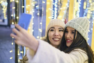 Two girlfriends asian girls in white coats are having fun hugging and selfie on mobile phone, new