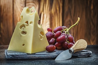Piece of maasdam cheese on vintage carving board knife and bunch of rose grape old wooden